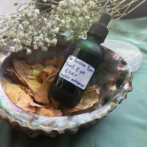 May include: A dark green glass bottle with a dropper, labeled "The Viridian Room Third Eye Elixir psychic enhancer," sits in an iridescent shell. The shell is filled with dried petals, and white flowers are in the background.