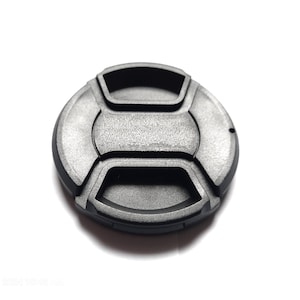 May include: A black camera lens cap with a textured surface. The cap is round with a series of raised, rectangular shapes around the edge and center. The cap is designed to protect the lens from damage.