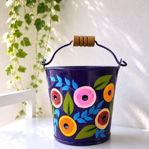 May include: A deep purple metal bucket featuring a hand-painted floral pattern with pink, orange, and yellow circular flowers accented by green and blue leaves. The bucket includes a matching purple metal handle with a cylindrical wooden grip.
