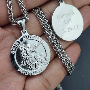 May include: A silver-colored Saint Michael pendant necklace with a detailed depiction of Saint Michael. The pendant is round with the words "SAINT MICHAEL PROTECT US". A second round pendant is engraved with the names "Gilbert & Lali" and the date "4-29-17".