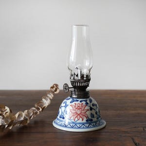 May include: A small oil lamp with a white porcelain base decorated with blue and red floral patterns. The lamp has a clear glass chimney and a silver metal top.