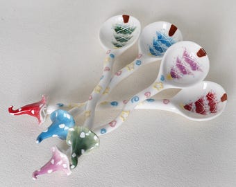 May include: Four white ceramic spoons featuring colorful painted Christmas tree designs in the bowls. The handles are decorated with small pastel stars, hearts, and swirls, ending in sculpted, pointed gnome-style hat finials in red, blue, green, and pink.