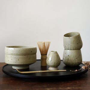 May include: A green ceramic matcha bowl with a wooden whisk, a small green ceramic cup, and three stacked green ceramic cups on a black tray.