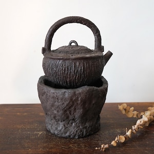 May include: A dark brown, textured cast iron teapot with a matching arched handle and a small spout, resting securely inside a heavy, rough-surfaced, dark brown cylindrical base. The teapot features a lid with a small central loop handle.