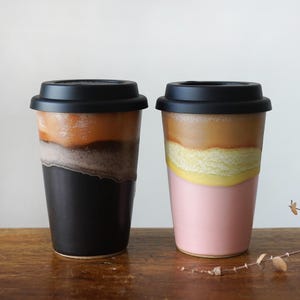 May include: Two ceramic travel mugs with black lids. One mug is black at the bottom, transitioning to gray, orange, and brown. The other mug is pink at the bottom, with yellow, tan, and brown layers. Both mugs are on a wooden surface.