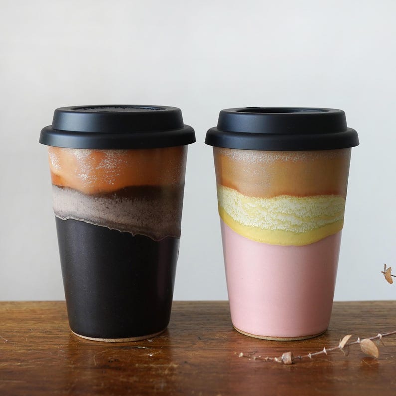 May include: Two ceramic travel mugs with black lids. One mug is black at the bottom, transitioning to gray, orange, and brown. The other mug is pink at the bottom, with yellow, tan, and brown layers. Both mugs are on a wooden surface.