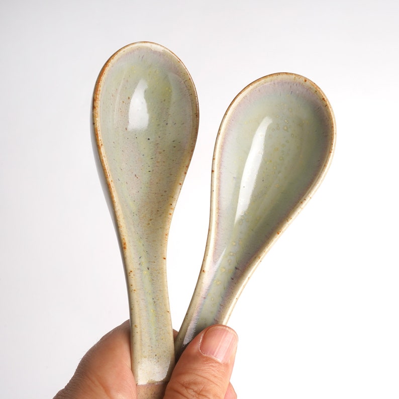 May include: Two ceramic soup spoons with a light green glaze and brown speckles. The spoons are held in a hand.