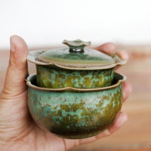 May include: A small, round ceramic tea bowl with a matching lid, featuring a scalloped rim and a mottled glaze in shades of deep forest green, olive, and earthy brown. The surface has a textured, speckled appearance with a glossy finish.