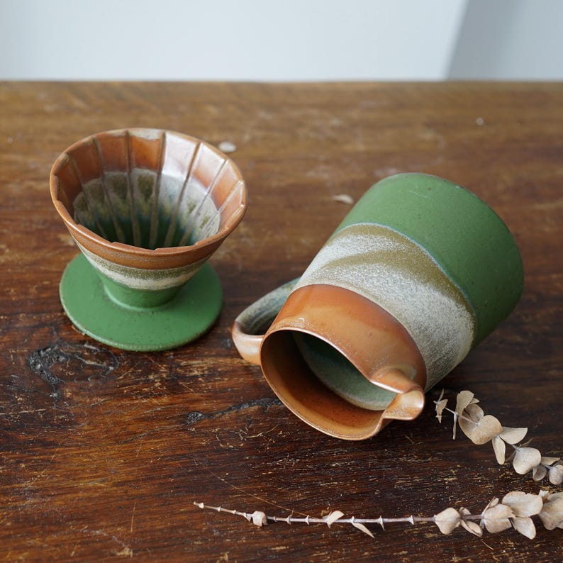 May include: A ceramic coffee pour-over and mug set. The pour-over is brown and green, with a ridged interior. The mug is green with a brown rim and handle. Both items have a rustic, handmade appearance, perfect for coffee lovers.