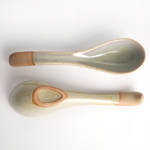May include: Two ceramic soup spoons with a speckled glaze in shades of beige and brown. The spoons have a rounded bowl and a long handle.