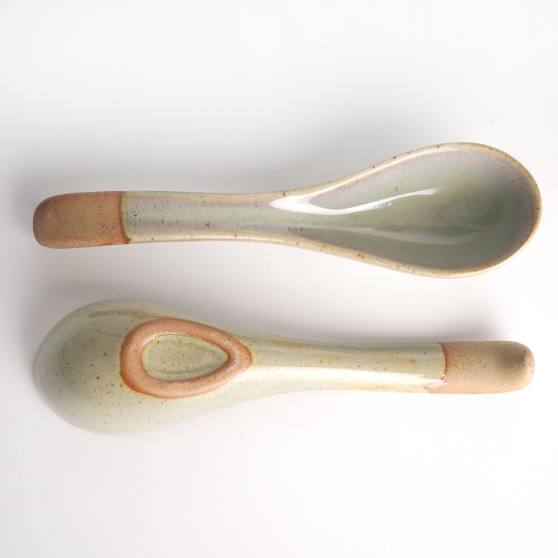 May include: Two ceramic soup spoons with a speckled glaze in shades of beige and brown. The spoons have a rounded bowl and a long handle.