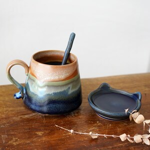 May include: A handmade ceramic mug with a matching lid and spoon. The mug features a unique color gradient, transitioning from tan to green and deep blue. The lid is dark blue and cat-shaped.