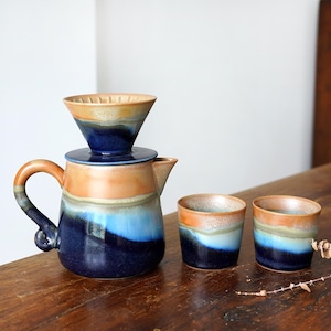 Op de afbeelding: Een keramische koffie-overgietset met een bijpassende karaf en twee kleine kopjes. De set heeft een kleurenpalet van blauw, oranje en bruin. De karaf heeft een handvat en een tuit. De kopjes zijn cilindrisch.