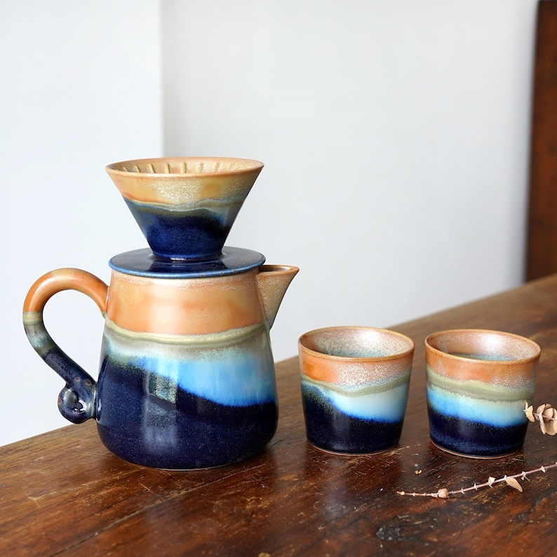 May include: A ceramic coffee pour-over set with a matching carafe and two small cups. The set features a color palette of blues, oranges, and tans. The carafe has a handle and a spout. The cups are cylindrical.