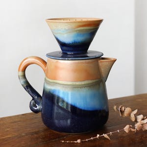 May include: A ceramic coffee pour-over set with a matching carafe and dripper. The set features a gradient of colors, including shades of blue, orange, and brown. The carafe has a curved handle and a spout for easy pouring. The dripper sits atop the carafe.