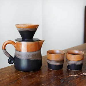 Puede incluir: Un juego de café de cerámica con un vertedor, una jarra y dos tazas a juego. El juego presenta un degradado de color negro, gris y naranja. La jarra tiene un asa curva y un pico. Las tazas son cilíndricas.