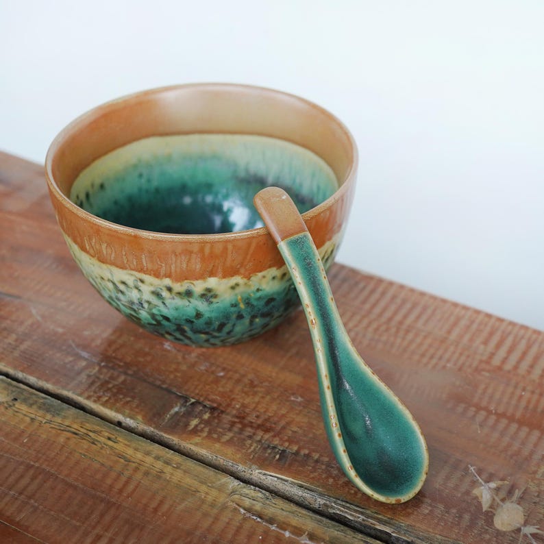May include: A ceramic bowl and matching spoon featuring a two-tone glaze. The upper portion of the bowl and the handle of the spoon are a warm tan color, while the lower section of the bowl and the spoon bowl feature a deep green mottled finish.