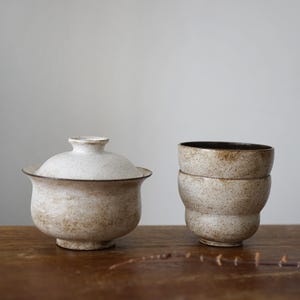 May include: A ceramic tea set featuring a lidded bowl and two stacked cups. The pieces have a speckled, off-white glaze with brown accents. The set is displayed on a wooden surface, suggesting a rustic or minimalist aesthetic.