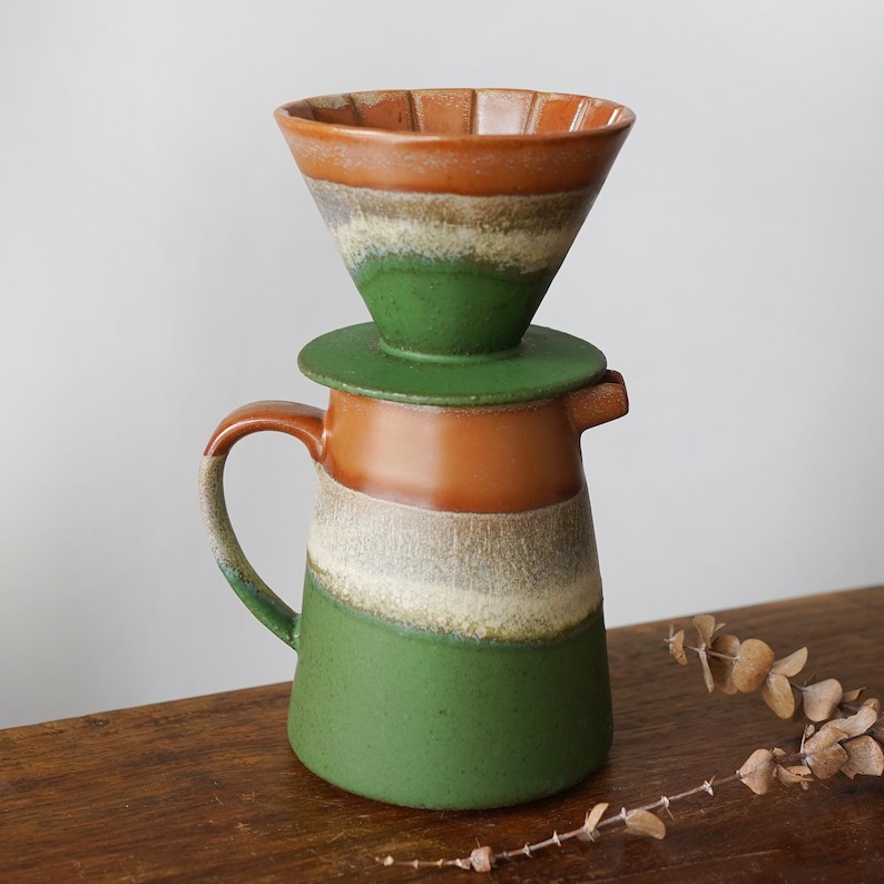 May include: A ceramic pour-over coffee maker set. The set includes a cone-shaped dripper, a lid, and a carafe. The dripper and carafe are in shades of brown, green, and white. The carafe has a handle and a spout.