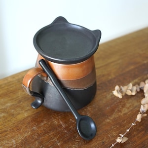 Könnte beinhalten: Eine Keramik-Tasse mit Deckel und Löffel. Die Tasse ist zylindrisch mit Henkel und mit horizontalen Bändern in Schwarz, Braun und Orange verziert. Der Deckel hat zwei kleine ohrförmige Vorsprünge. Ein schwarzer Löffel liegt an der Tasse.