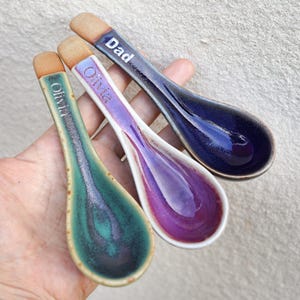 May include: Three ceramic spoons with glazed bowls in teal, purple, and dark blue. Each handle features a natural tan unglazed tip and is personalized with white text: two spoons are inscribed with the name Olivia and one with the word Dad.
