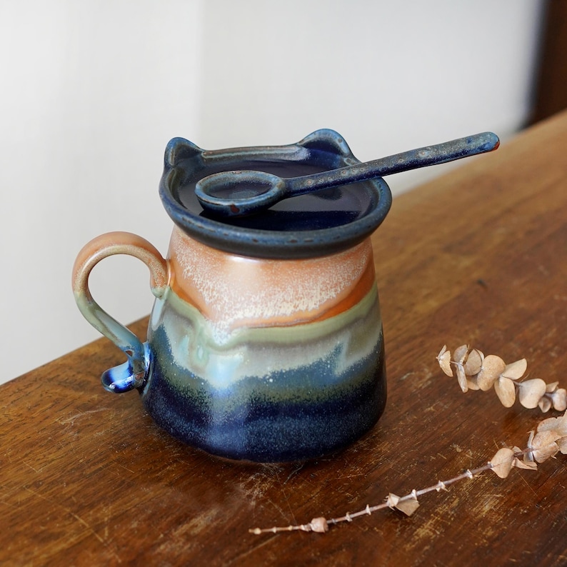 May include: A ceramic mug with a cat-ear lid and matching spoon. The mug features a color gradient of blue, green, orange, and tan. The handle and spoon are blue. The mug is sitting on a wooden surface.