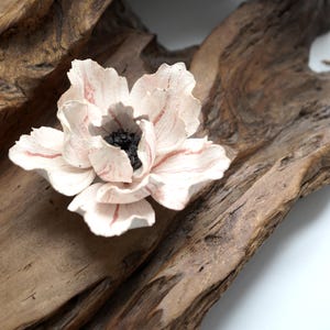 Op de afbeelding: Een handgemaakte, lichtroze bloem met rode aderen en een zwart centrum, rustend op een stuk verweerd, bruin drijfhout. De bloemblaadjes zijn fijn gedetailleerd, wat een natuurlijke, artistieke esthetiek creëert.