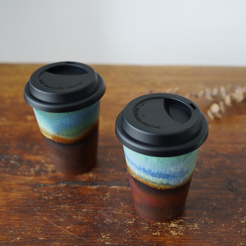 May include: Two ceramic to-go mugs with black lids. The mugs have a blue, green, and brown glaze with a layered effect.