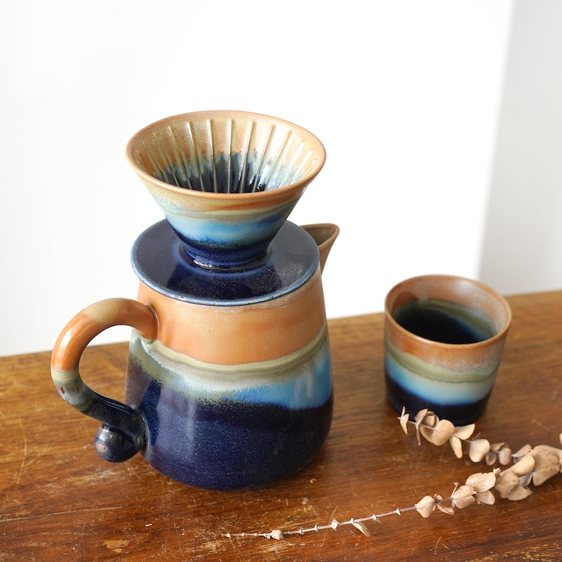 May include: A ceramic pour-over coffee maker set with a matching cup. The coffee maker is a combination of blue, orange, and brown hues. The cup and coffee maker are on a wooden surface.