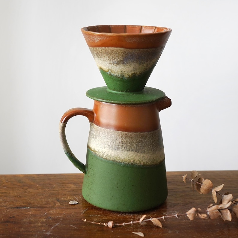 May include: A ceramic coffee pour-over set with a brown, green, and cream color scheme. The set includes a cone-shaped dripper, a lid, and a pitcher with a handle. The design is rustic and handmade, perfect for brewing coffee.