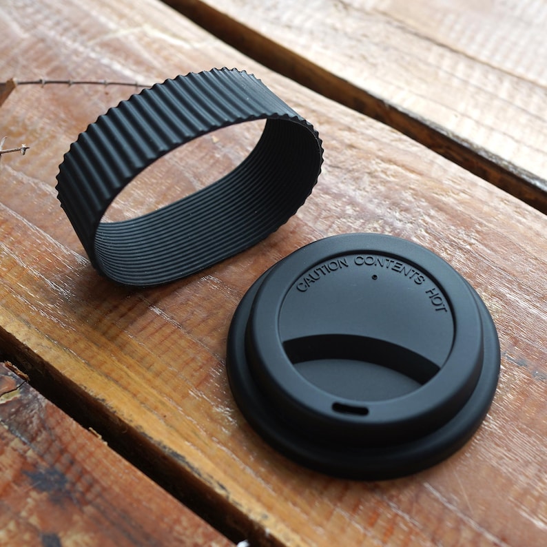 May include: Black coffee cup lid and sleeve set. The lid has the words "CAUTION CONTENTS HOT" printed on it. The sleeve is a ribbed, flexible band. Both items are made of a matte black material and are displayed on a wooden surface.