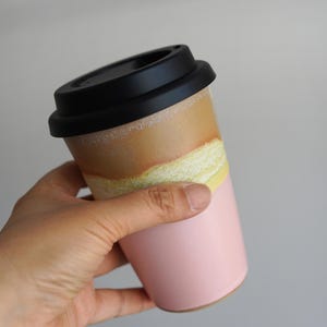 May include: A ceramic travel mug with a black lid. The mug is pink at the bottom, transitioning to yellow and brown at the top. The mug is held in a hand against a neutral background.