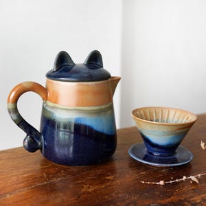 May include: A ceramic teapot and pour-over coffee maker set. The teapot is navy blue with orange and blue accents, and cat ear-shaped lid. The coffee maker is a matching color scheme. The set is on a wooden surface.