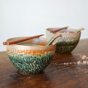 May include: Two ceramic bowls with a reactive glaze featuring a gradient of earthy brown at the rim transitioning into a textured, mottled deep green and black base. Each bowl contains a pair of wooden chopsticks resting across the top opening.