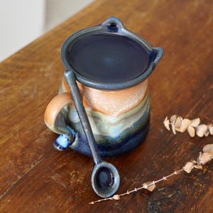 May include: A handcrafted ceramic mug and spoon set. The mug has a dark blue interior and rim, with a multi-coloured exterior of orange, green, and blue. The spoon is placed beside the mug, forming a coordinated ensemble.