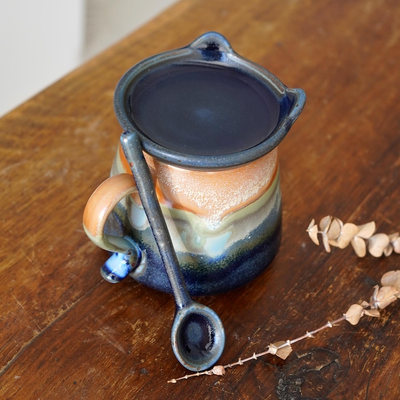 May include: A handmade ceramic mug with a matching spoon. The mug features a dark blue interior and rim, with a colorful exterior of orange, green, and blue hues. The spoon rests on the side of the mug, creating a cohesive set.