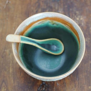 May include: A ceramic bowl with a deep teal interior and a light beige exterior rim, featuring a matching ceramic spoon resting inside. The spoon has a teal bowl and a light beige handle, complementing the bowl's dual-tone color scheme.