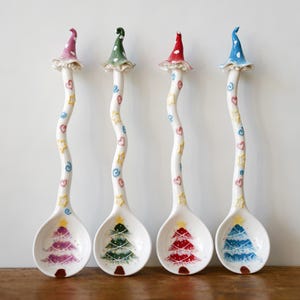 May include: Four decorative ceramic spoons with white, wavy handles decorated with hearts, stars, and swirls. Each spoon has a Christmas tree design in the bowl and a hat-shaped topper in pink, green, red, and blue. The spoons are for decorative use.