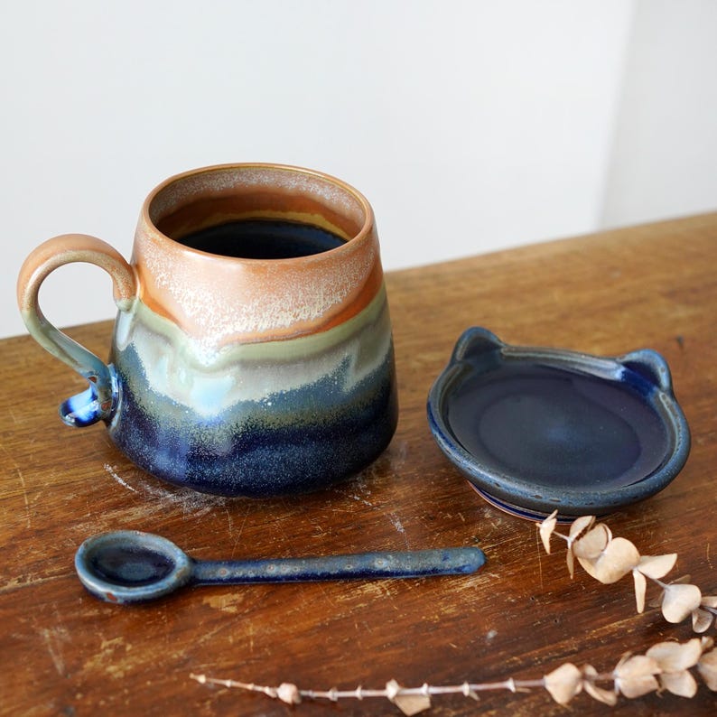 May include: A ceramic mug with a matching cat-shaped lid and spoon. The mug features a gradient of colors, including blue, green, orange, and brown. The spoon and lid are a deep blue. The set is placed on a wooden surface.