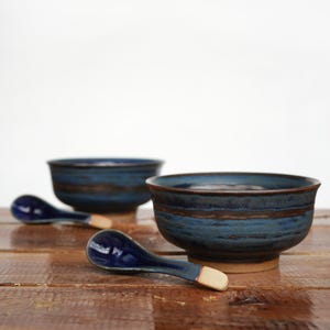 Könnte beinhalten: Zwei blaue Keramikschalen mit braunem Band und passenden Löffeln. Die Schalen stehen auf einer Holzoberfläche. Die Löffel haben eine blaue Schale und einen beigefarbenen Griff. Die Schalen haben einen Durchmesser von ca. 13 cm.