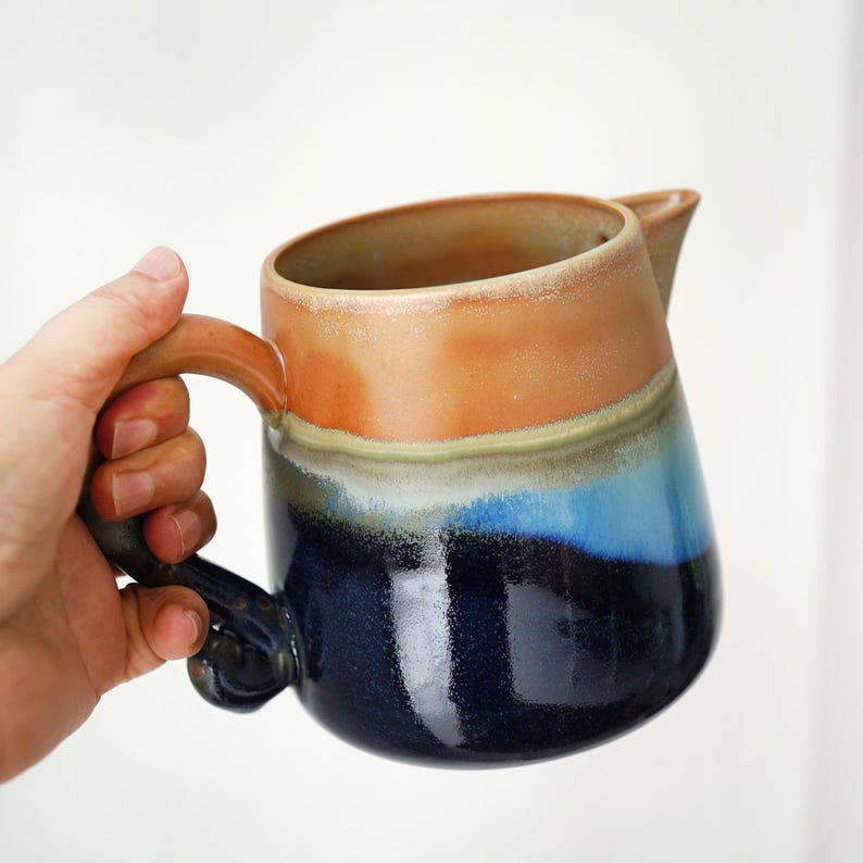 May include: A handmade ceramic pitcher with a unique design. The pitcher features a gradient of colors, including shades of brown, blue, and navy. It has a curved handle and a spout for pouring. The pitcher is held by a hand against a white background.