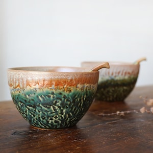 Pode incluir: Duas tigelas de cerâmica com acabamento texturizado e esmaltado. As tigelas apresentam um gradiente de cores, incluindo verde, laranja e castanho. As tigelas estão sobre uma superfície de madeira. Uma tigela tem uma colher de madeira.
