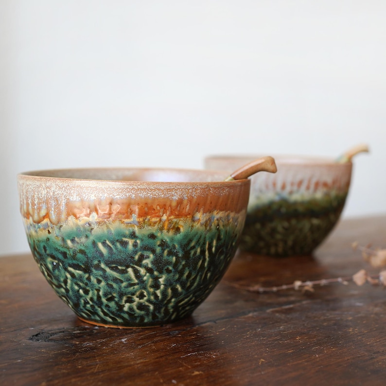 May include: Two ceramic bowls with a textured, mottled glaze featuring a deep forest green base that transitions into a warm, earthy brown rim. Each bowl has a rounded shape and a thick, uneven rim, with a small matching ceramic spoon resting inside.