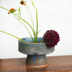Op de afbeelding: Een keramische voetvaas met een gespikkeld blauw en grijs exterieur, met groene stelen en oranje en bordeauxrode bloemen. De vaas heeft een brede, ondiepe kom en een stevige voet, ideaal voor bloemstukken.