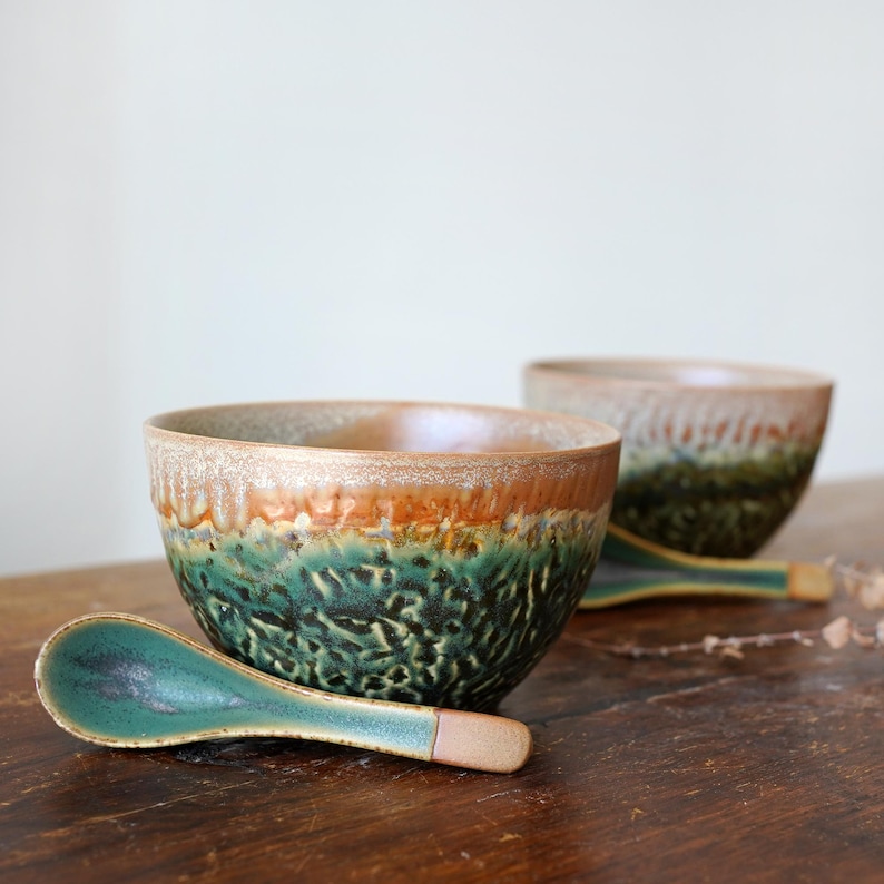 May include: Two ceramic bowls and two matching spoons featuring a reactive glaze with a textured, dark green and black base that transitions into a smooth, light brown rim. The bowls have a rounded shape and the spoons have a long, curved handle.
