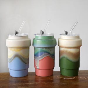 Könnte beinhalten: Drei Keramikbecher mit Deckeln und Strohhalmen. Jeder Becher hat ein einzigartiges Farbschema, darunter Blau, Grün und Braun, mit einem geschichteten, strukturierten Design. Die Becher sind für kalte Getränke konzipiert.