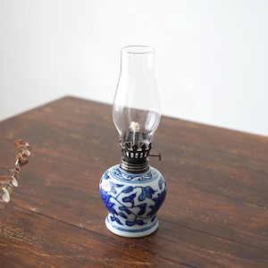 Pode incluir: Uma pequena lâmpada de óleo de porcelana azul e branca com uma chaminé de vidro transparente. A lâmpada tem um design floral e está sentada em uma mesa de madeira.
