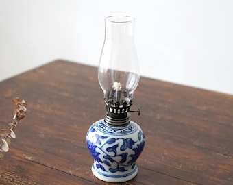 May include: A small blue and white porcelain oil lamp with a clear glass chimney. The lamp has a floral design and is sitting on a wooden table.