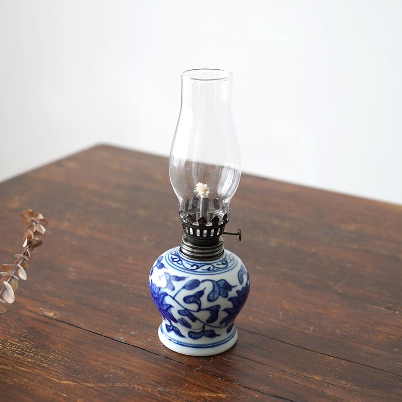 Puede incluir: Una peque&ntilde;a l&aacute;mpara de aceite de porcelana azul y blanca con una chimenea de vidrio transparente. La l&aacute;mpara tiene un dise&ntilde;o floral y est&aacute; sentada sobre una mesa de madera.