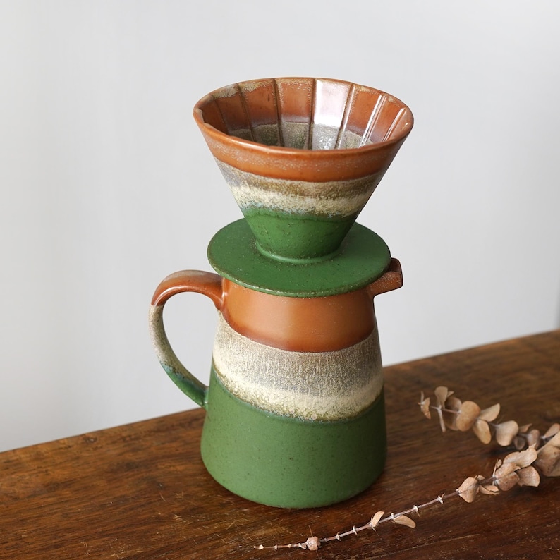 May include: A ceramic pour-over coffee maker with a brown, green, and white color scheme. The coffee maker includes a cone-shaped filter holder and a matching carafe. The design features a rustic, handcrafted aesthetic, perfect for brewing coffee.